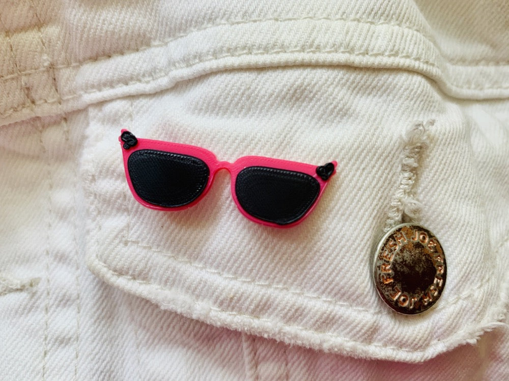 There is a close up of a white demin jacket pocket. An R+D pin is attached that looks like a pair of cat eye sunglasses. They are hot pink with black frames and black accents in the corners. 