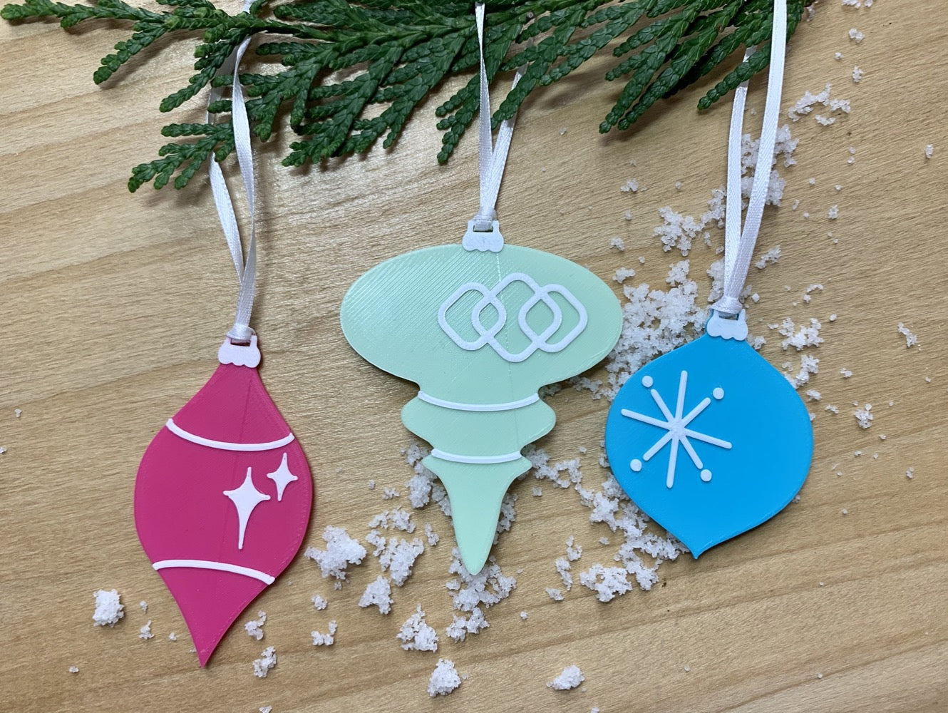 On a wood background with white snow and hanging from an every green branch are a set of 3 R+D 3D printed ornaments. They are all designed to look like vintage baubles that would have commonly been found on Christmas trees in the past. They each have white accents of stars and shapes. One is hot pink, one is mint green, and one is teal.