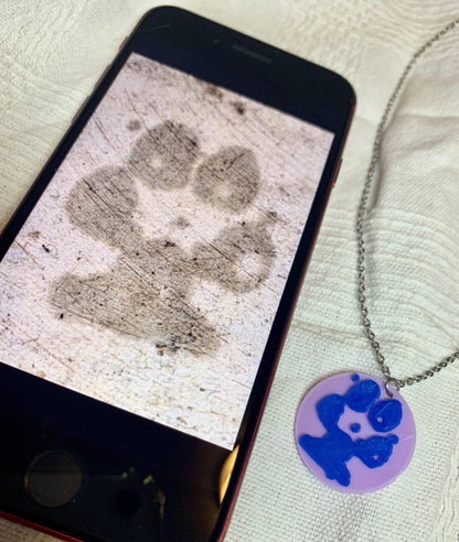 Shown laying on a white fabric is a phone and a necklace with a 3D pritned pendant. The pendant is a 1 inch light purple circle with a bright blue paw print on it. The picture on the phone is of the same paw print and was used to create the custom print. 