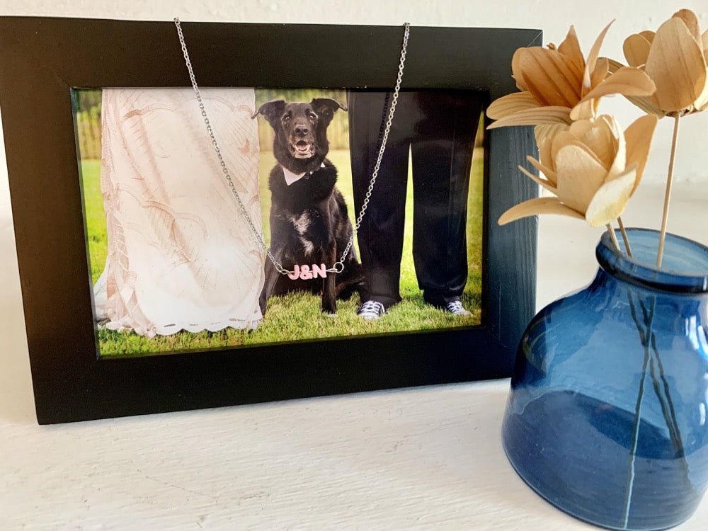 Next to a vase is a frame with a picture of a bride and groom with a dog sitting between them. Draped over the frame is a necklace with small letters on a metal bar. The letters are 3D printed like beads and will rotate and move around. This necklace has 3 characters (J&N) in a light pink color. 