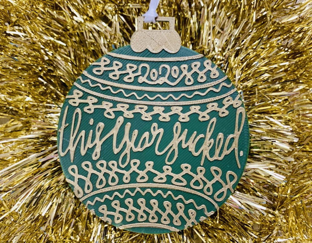 On a background of bright gold tinsel garland is a R+D 3D printed ornament. The ornament is shaped like a traditional bulb. It is dark green with gold drawings and script on it. Worked into the doodles are the year 2020 and then the words, "this year sucked".