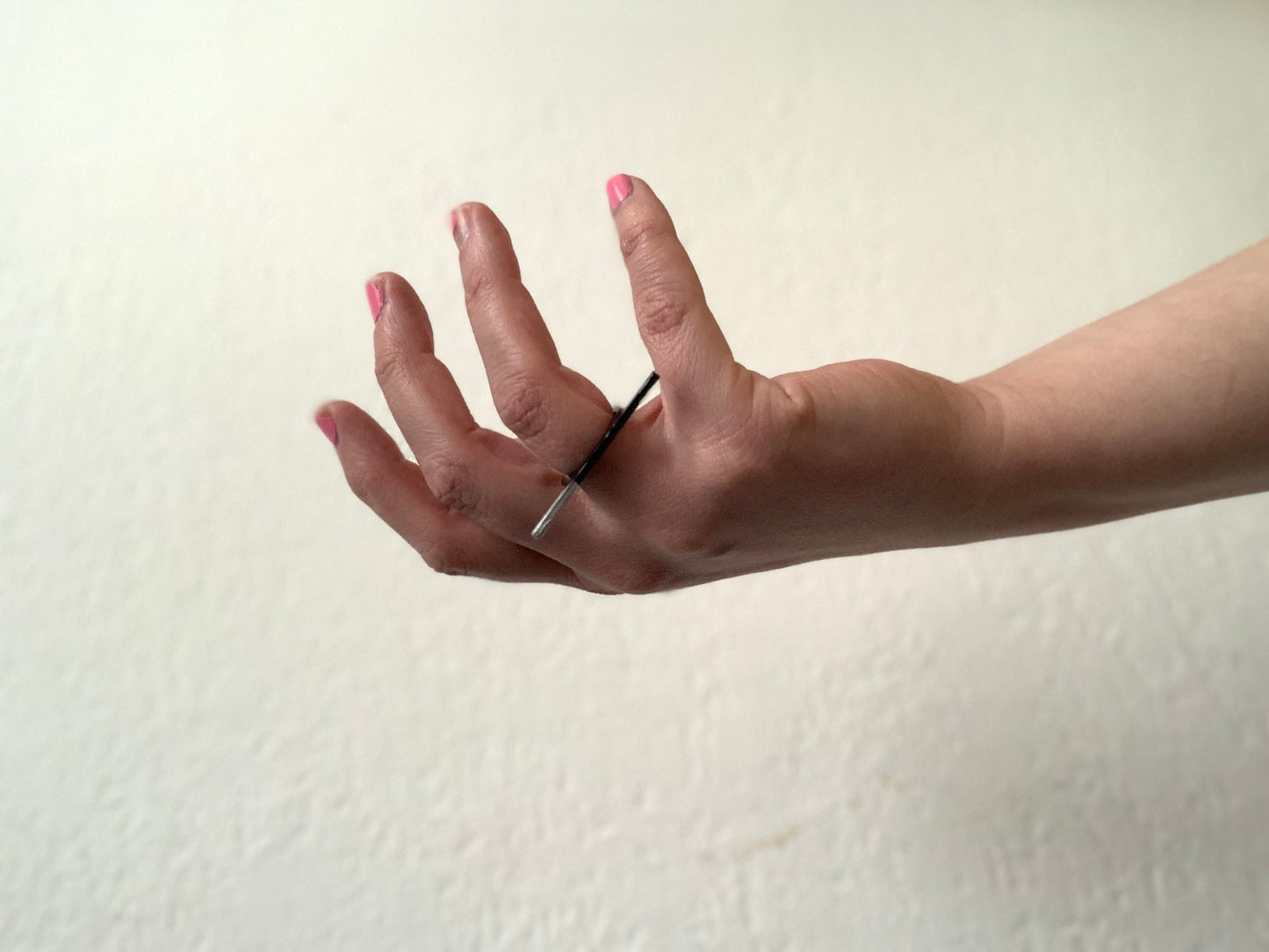 A hand is reaching into the frame across a white background. The hand is turned up like it may be recieving something. On the ring finger is a 3D printed ring that has a bar across the top. The bar is dipped in metallic paint on one side and extends over the middle and pinky fingers. 