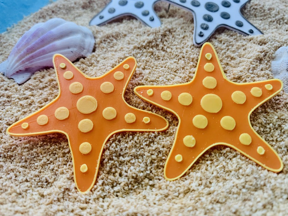 In the foreground are two R+D earrings. They are shaped like star fish and are orange with yellow outlines. There are yellow circles that stretch out on each leg of the starfish. They are laying on sand and shells and other star fish earrings are visible in the background. 