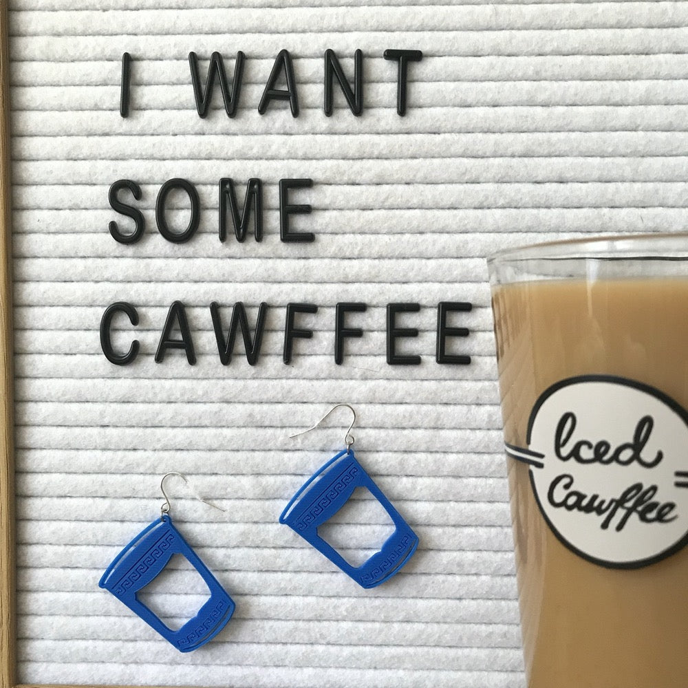 There is a white felt letter board with black letters reading, I want some cawffee and two earrings hanging below the words. The earrings look like classic Greek to-go coffee cups from New York City diners and bodegas. To the side of the letter board and earrings is a pint glass filled with milk and iced coffee from Fish's Eddy. It reads , Iced Cawffee in scripted lettering.