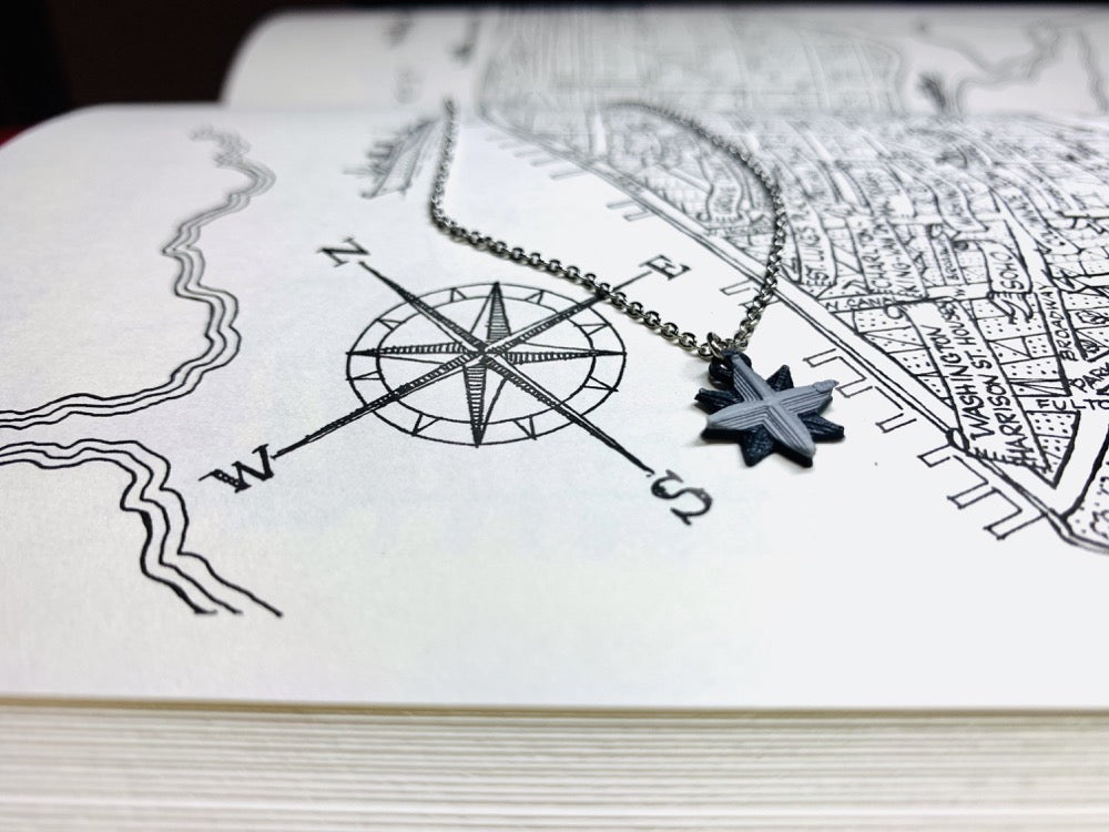 A book is opened to a drawn map of Manhattan, NY. Visable is the compass in the corner indicating North, East, South, and West. Laying next to the compass is an R+D necklace. The necklace is small and detailed. It is shaped like a compass and 3D printed in black and silver. 