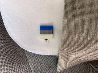 A close up of the sleeve of a grey suit reveals a white french cuff with a 3D printed cufflink. The cufflink is square with blue, silver and white stripes to match the Sunnyside Arch lapel pin/tie tack.