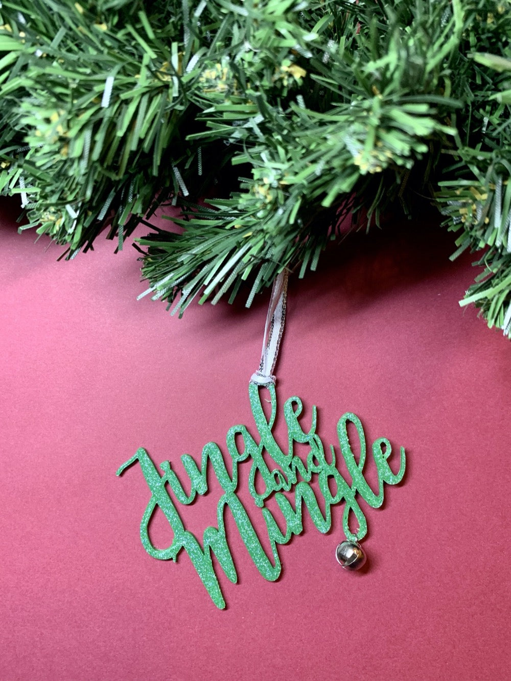 On a bright red background and hanging from a green wreath is a 3D printed R+D ornament. It is a cursive text with a jingle bell and covered in glitter to make it shimmer and shine in the light. This ornament reads, Jingle and Mingle.