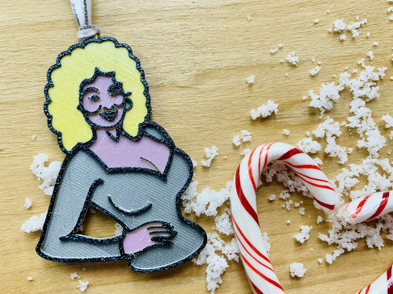 On a wood background with white snow and red and white candy canes is a R+D 3D printed ornament. This ornament is shaped like Dolly Parton. You can see one hand resting on her hip as she wears a silver top. Her trademark neckline and big blond hair is also visible with a smile. The entire ornament is covered in glitter to be able to shimmer and shine in the light.