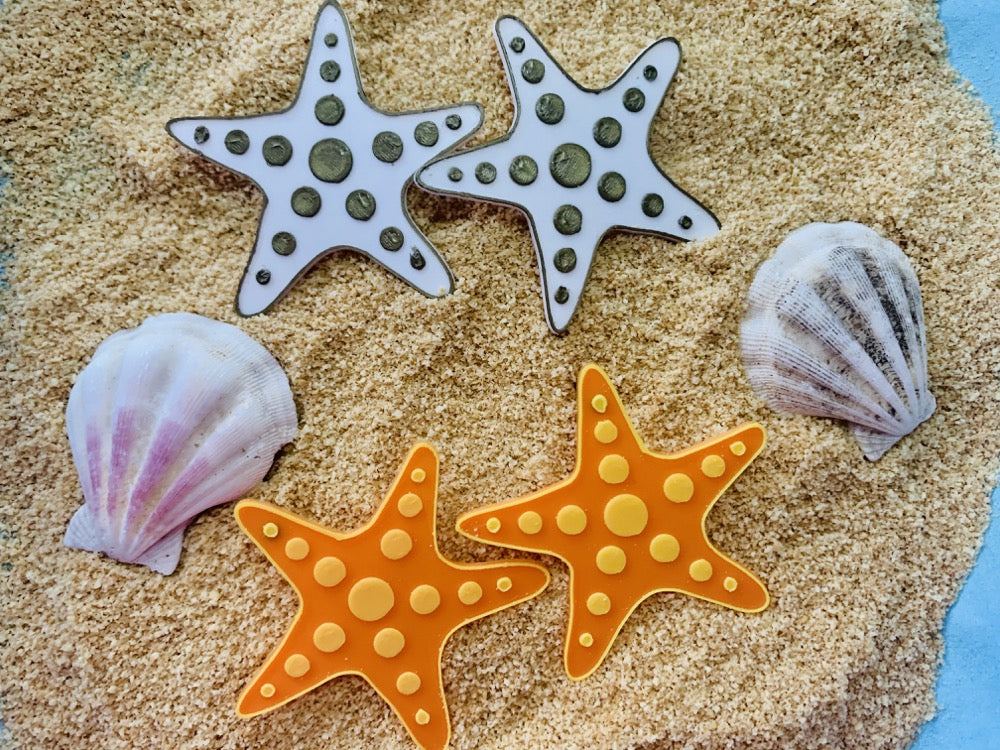 On a bed of sand are sea shells and two sets of earrings. The earrings are both shaped like star fish, with circles that are stretching out across each of the arms. One of the earring sets is gold and white, the other is orange and yellow. 