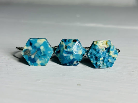 There are three rings side by side on a white background. They each have a hexagon shape with a twisted metal band. They are made from recycled remnants of plant based filament which gives them a speckled look like granite. The first ring has a smooth surface on the top, the middle a faceted texture and the third a rough texture like a geode being formed. These rings are all using teal, blue, white, and black filaments to make these one of a kind pieces.