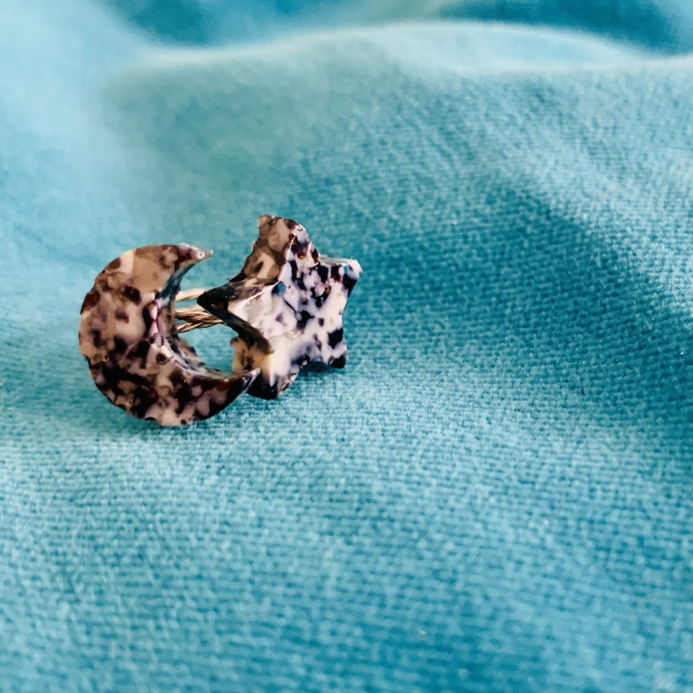 On a teal velvet pillow are two rings next to each other. One is a cresent moon shape, the other a star. They are cast from recycled 3D prints in black, white and silver plant based filaments. They have a speckled look like granite.