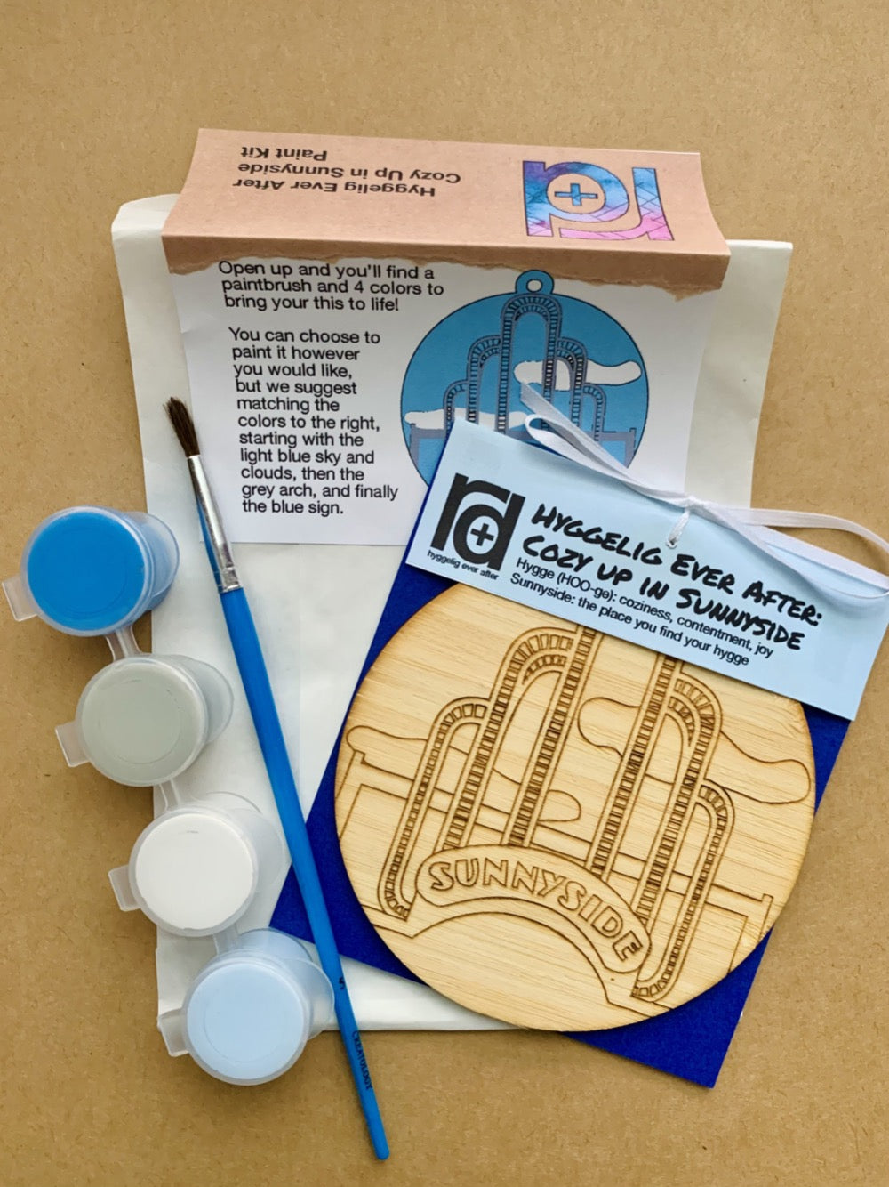 Shown laying on craft paper is the packaging and contents of this DIY Paint Kit. There is a paper envelope with instructions, a paintbrush, a set of 4 paint wells and a wall hanging with a laser cut NYC landmark.