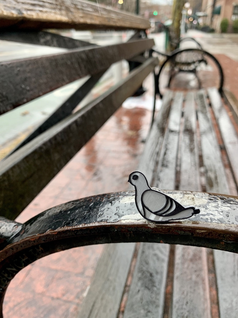 Resting on the arm of a park bench is a close up of a 3D printed pin. It is silver and black, shaped like a pigeon and 3D printed with plant based materials. 