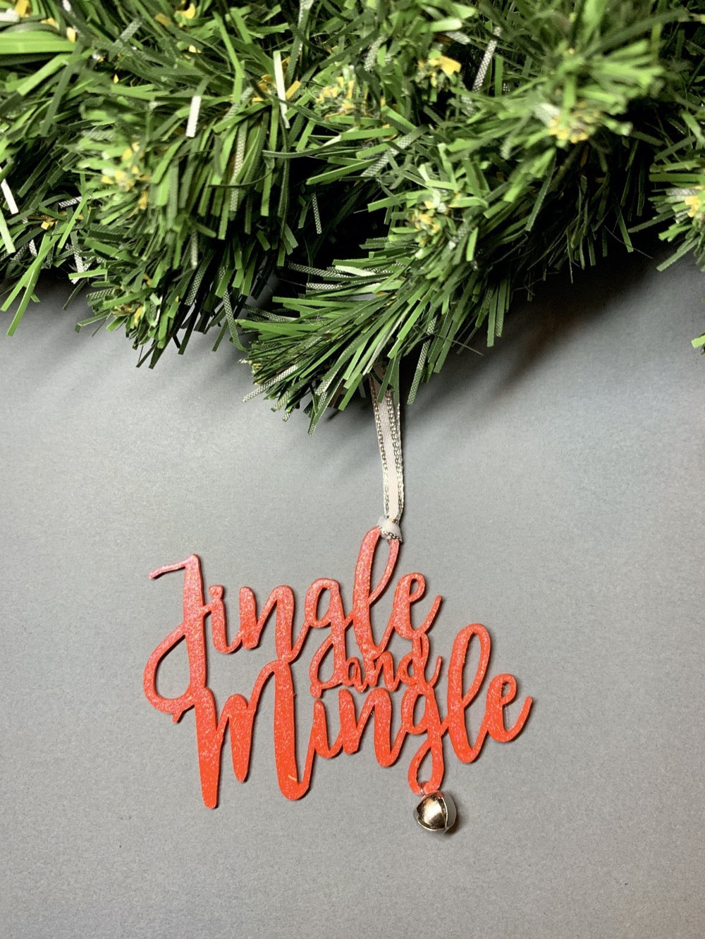 On a grey background and hanging from a green wreath is a 3D printed R+D ornament. It is a cursive text with a jingle bell and covered in glitter to make it shimmer and shine in the light. This ornament reads, Jingle and Mingle.