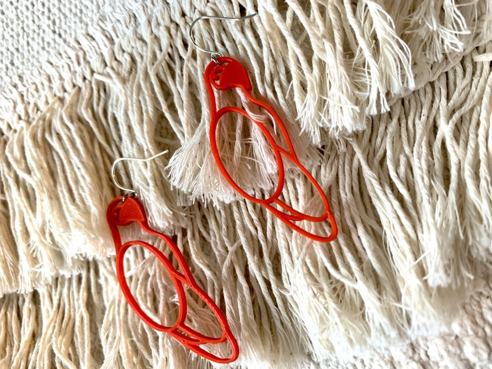 Two R+D earrings are laying on a background of cream tassels. The earrings are bright red and shaped like parrots. 