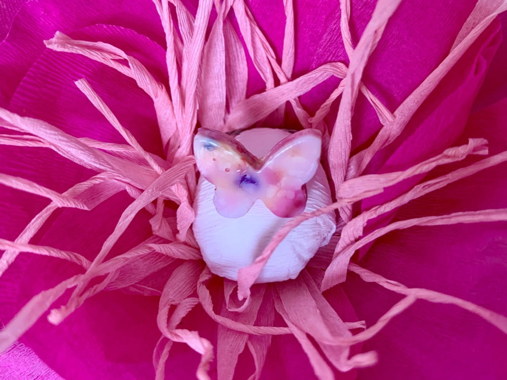 In the center of a paper anemone flower is a butterlfy ring. The ring is cast from recycled 3D prints in shades of pink, purple, yellow, orange, and white. It has the appearance of granite or marble. 