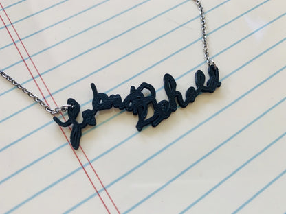 Shown on a ceramic board with lines like a piece of notebook paper is a 3D printed necklace. The pendant on the necklace is based off of the handwriting in an old card.