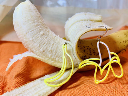 On an orange cloth napkin there is a banana that is partially peeled open. Hanging off of the peel is an R+D earring that is also a half peeled banana. It is yellow and reveals a white banana. Resting on another peel is a second R+D earring that is the shape of an unpeeled banana. 