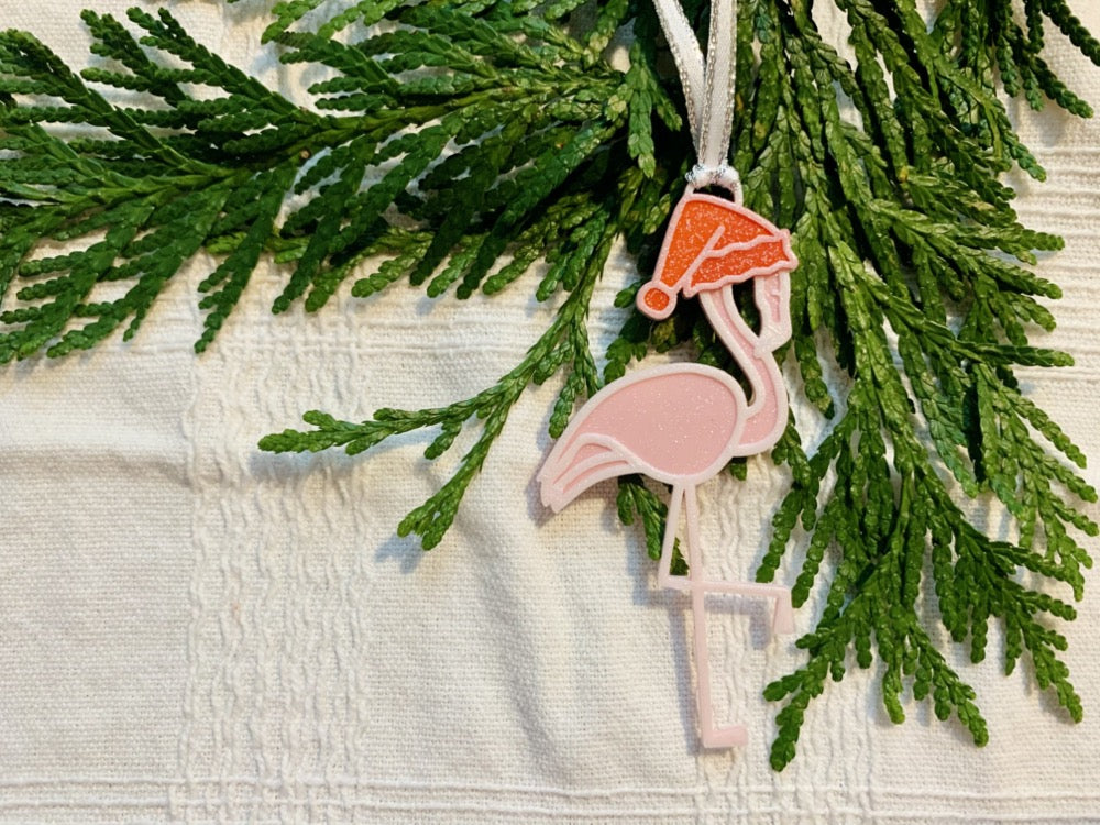 On a white fabric background with sprigs of evergreen is a R+D 3D printed ornament. It is a light pink flamingo that has one leg up and is wearing a red santa hat.
