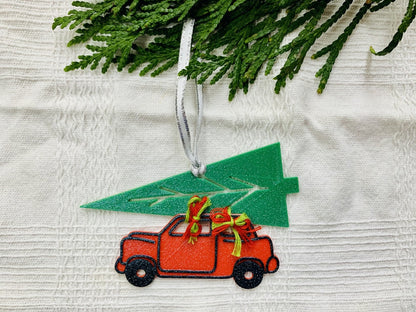On a white fabric background with a branch of evergreens at the top there is a R+D 3D printed ornament hanging down. It is shaped as vintage style red car with a big green tree strapped on top. It is held together with red and green bows. The entire ornament is covered in glitter to be able to shimmer and shine in the light. 