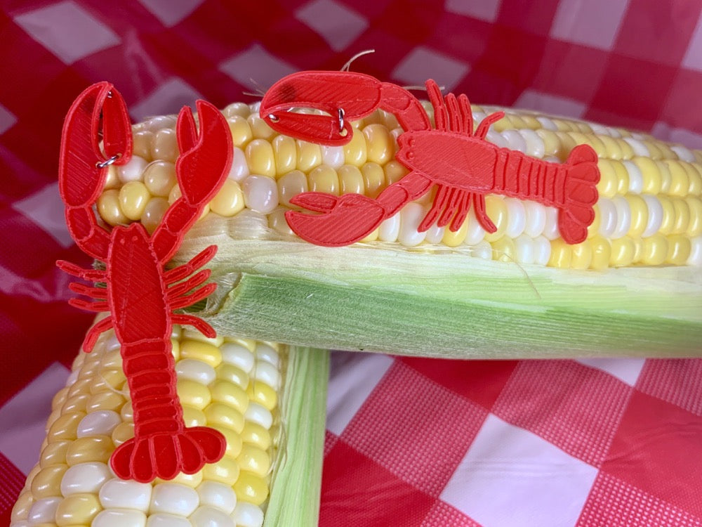 In the far background is a checkered white and red tablecloth from a classic picnic setting. In the foreground are two cobs of corn that are partially shucked. Resting on those cobs are two 3D printed earrings that are shaped like lobsters. They are bright red. 