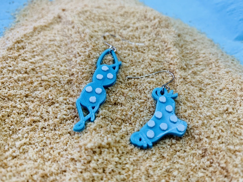 On a light blue background is a pile of fluffy sand. There are two earrings in the center. They are asymmetrical  earrings  one is a bikini top the other is a bikini bottom. They are both teal with white polka dots.