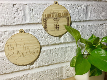 Shown on a white brick wall with a houseplant in the corner are two laser cut wall hangings. They are each cut from bamboo, a fast growing and sustainable grass. Etched on one is a picture of the Gantries in Long Island City, NYC. These gantries are an icon of the area and looked on from Manhattan and Gantry State Park. The other shows the nyc skyline with the statue of liberty, empire state building, Chrysler building and brooklyn bridge all visible.
