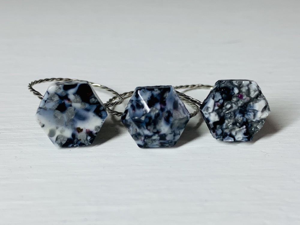 There are three rings side by side on a white background. They each have a hexagon shape with a twisted metal band. They are made from recycled remnants of plant based filament which gives them a  speckled look like granite. The first ring has a smooth surface on the top, the middle a faceted texture and the third a rough texture like a geode being formed. These rings are all using black, silver and white filaments to make these one of a kind pieces. 