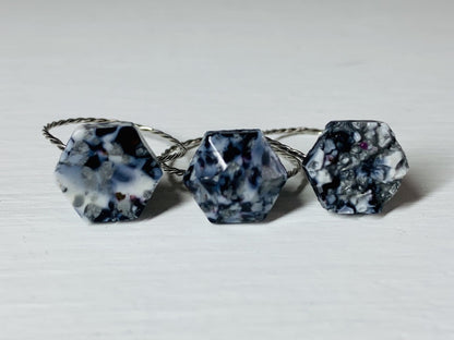 There are three rings side by side on a white background. They each have a hexagon shape with a twisted metal band. They are made from recycled remnants of plant based filament which gives them a  speckled look like granite. The first ring has a smooth surface on the top, the middle a faceted texture and the third a rough texture like a geode being formed. These rings are all using black, silver and white filaments to make these one of a kind pieces. 