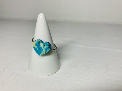 On a white background and slipped on a white cone ring holder is a ring that is cast from recycled 3D prints. The ring is in a heart shape with teal, blue, white, and black colors intermixed to be speckled like granite.