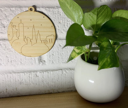 Shown on a white brick wall and next to a house plant is a laser cut wall hanging. It is cut from bamboo, a fast growing and sustainable grass. Etched in it a picture of the New York City Skyline with the Statue of Liberty, Empire State Building, Chrysler Building and Brooklyn Bridge all visible. Each piece can be painted like a paint by number or left plain. It can hang from your Christmas Tree or on you wall all year round.