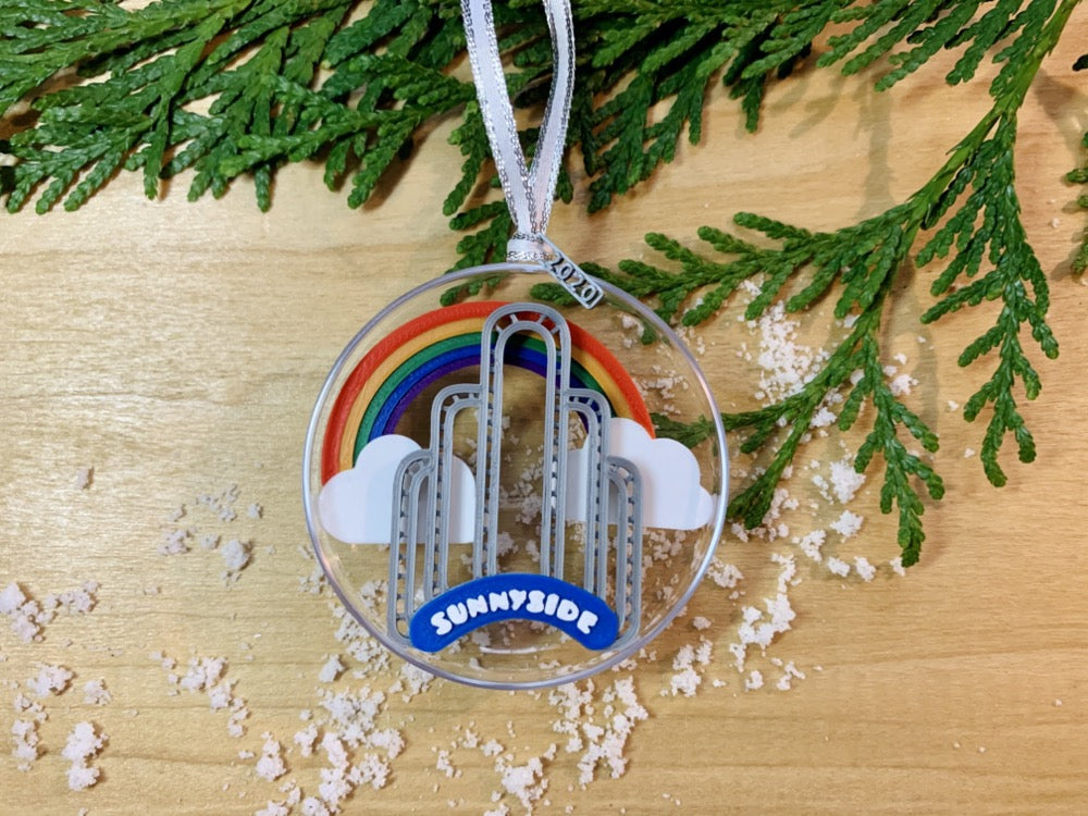 On a wooden shelf, there is a sprig of greenery and some scattered snow.Laying on top is a ornament with a small tag that has been 3D printed to read 2020. Inside the half circle ornament is a rainbow spanning between two clouds and the iconic Sunnyside Arch in front of it. 