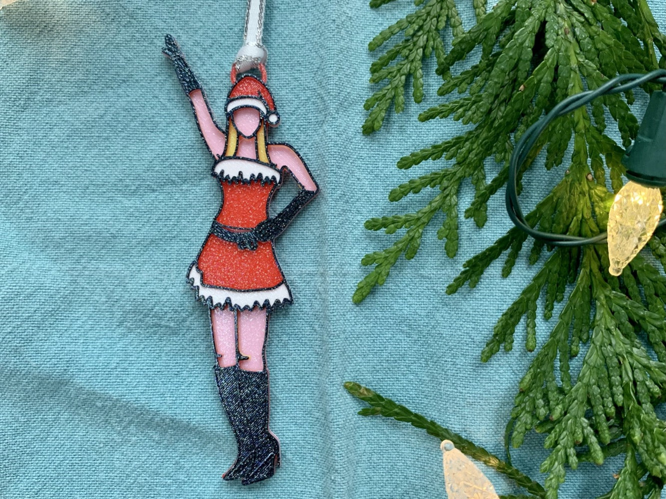 On a bright blue fabric background with green branches and holiday lights to the side is a 3D printed ornament from R+D. The ornament is printed in a plant based filament. It is shaped like Regina George from the movie Mean Girls. She is striking the iconic pose at the beginning of performing Jingle Bell Rock, wearing black gloves and boots and a red outfit with white trim. The entire ornament is covered in glitter to shimmer and shine in the light.