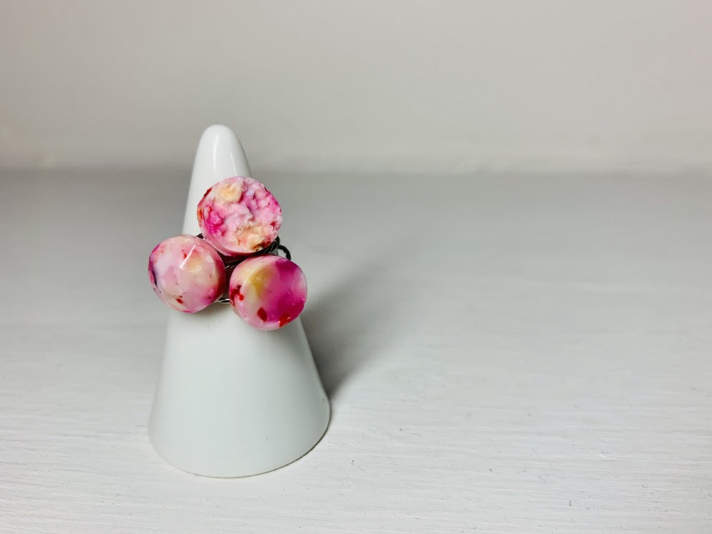 On a white background is a white ceramic ring holder with three cast rings on it. The rings all have a circular shape: one is a bumpy geode, another is a faceted circle, and finally one is a smooth circle. The rings are cast from recycled 3D prints in warm colors like shades of pink, purples, yellow, orange, red and white. They have the appearance of marble or granite. 