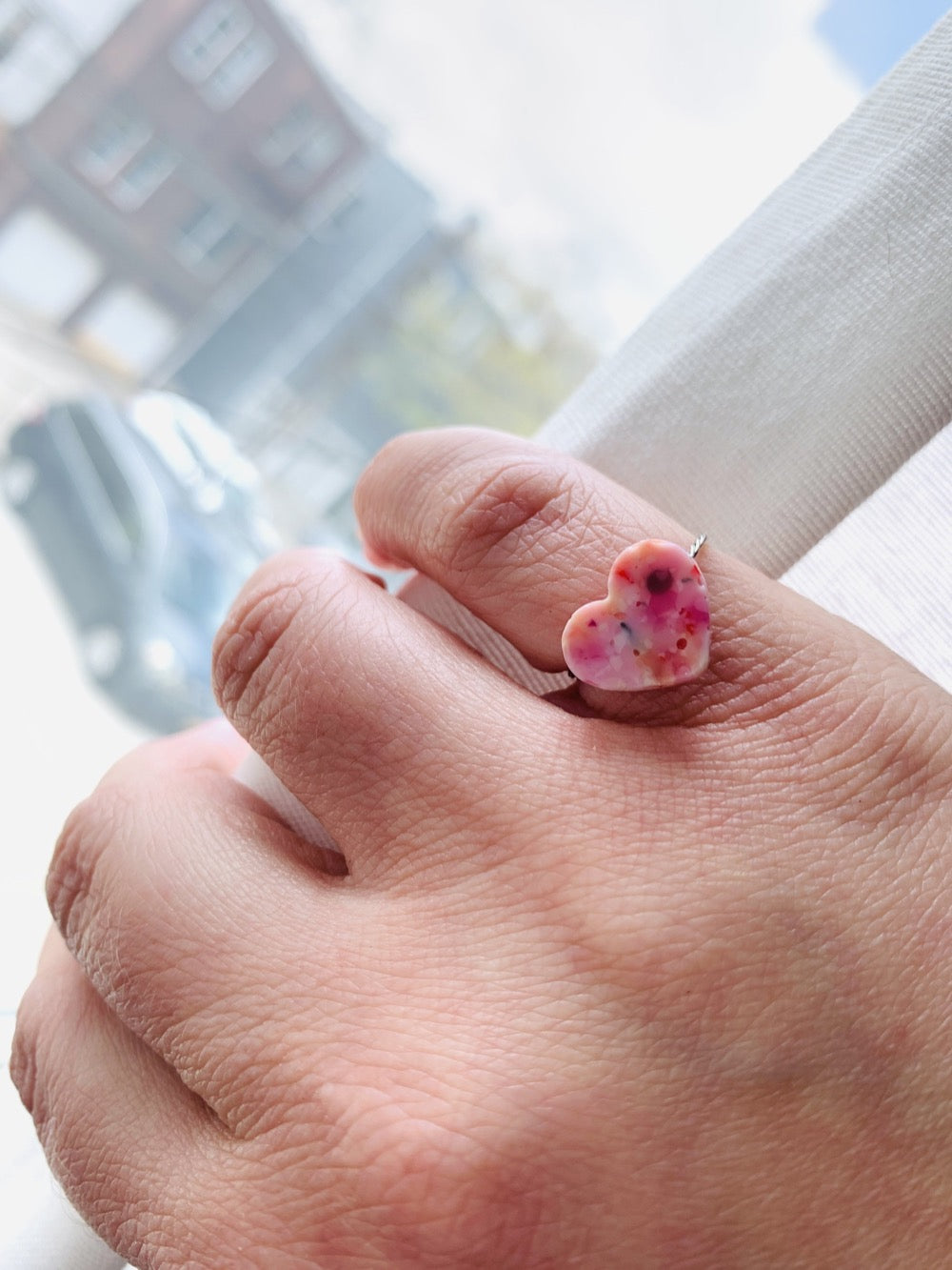 A hand is pulling back a white curtain to reveal a bright outside. On their pinky finger there is a ring with a heart charm. The charm has a smooth surface and is cast from recycled 3D prints in pinks, oranges, reds, yellows, and whites. 