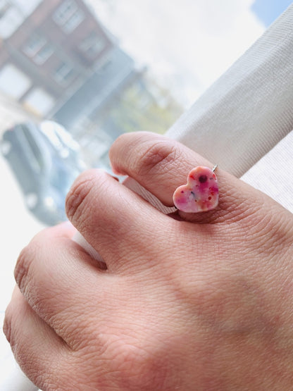 A hand is pulling back a white curtain to reveal a bright outside. On their pinky finger there is a ring with a heart charm. The charm has a smooth surface and is cast from recycled 3D prints in pinks, oranges, reds, yellows, and whites. 