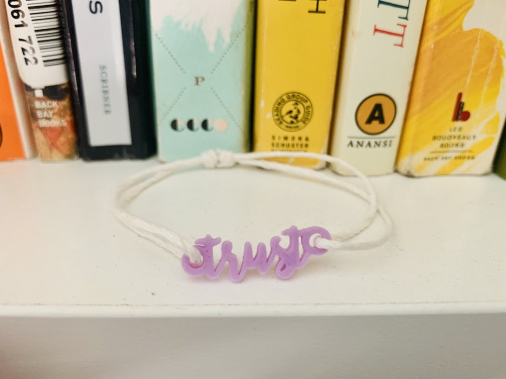 On a bookshelf ledge is a bracelet with an adjustable cotton cord. In the middle is a light purple 3D printed charm that says trust.