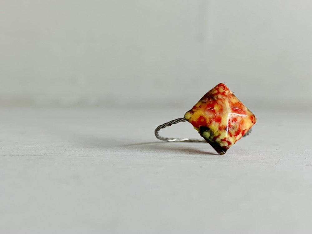 On a white background is a ring that is cast from recycled 3D prints. The ring is in a faceted square shape with red, yellow, orange, and black colors intermixed to be speckled like granite.