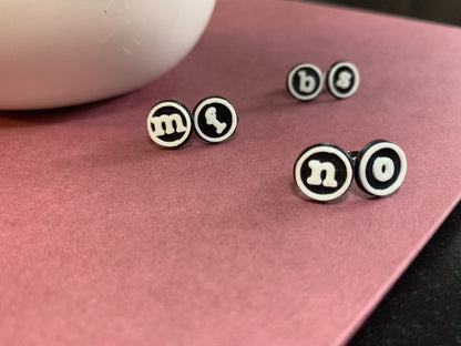There are three pairs of earrings shown. These r+d earrings are shaped like typewriter studs, with black backgrounds, white circle outlines and a lowercase letter in the center. They are 3d pritned with an eco friendly and plant based material. The earring pairs are "bs", "mf", and " no". They are resting on a muted red book with a white ceramic planter next to it. 