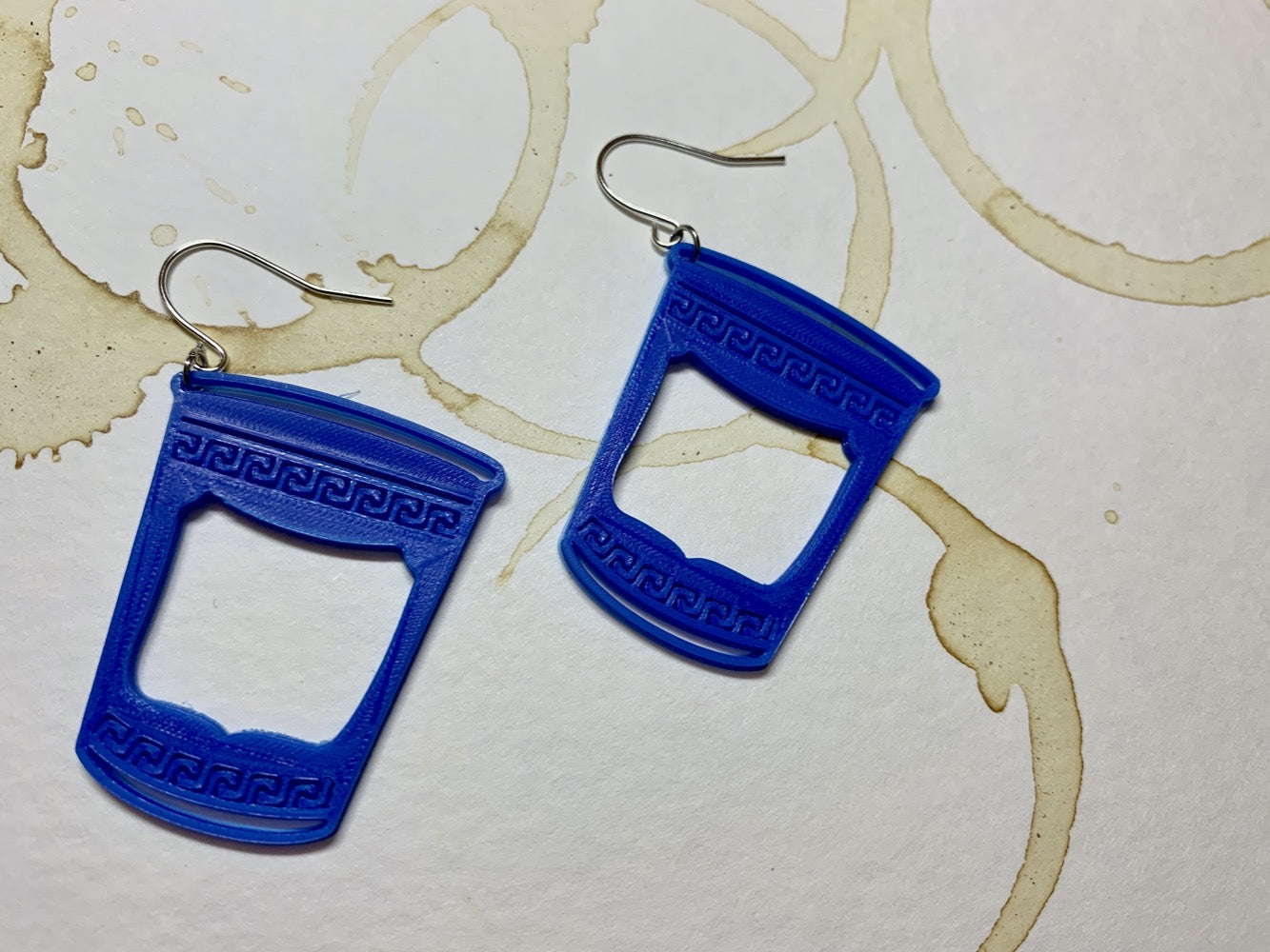 Two earrings that are made of a plant based plastic. They are in the shape of to-go coffee cups: The iconic ones from Greek diners or NYC bodegas with swirling borders and a big panel in the center. These earrings are resting on a paper that is stained with coffee rings overlapping one another. 