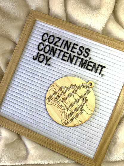 Shown on a cozy blanket is a letter board that reads "Coziness, Contentment, Joy." There is also a laser cut wall hanging. It is cut from bamboo, a fast growing and sustainable grass. Etched in it a picture of the iconic Sunnyside Arch in the center of the Queens neighborhood. Each piece can be painted like a paint by number or left plain. It can hang from your Christmas Tree or on you wall all year round.