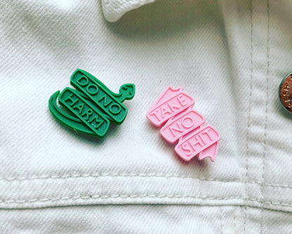 On a white denim jacket are two R+D pins. One pin is a bright kelly green that is a snake formed into a banner and reads do no harm. The other is a light pink banner that reads take no shit. They are both 3D printed with a plant based filament.
