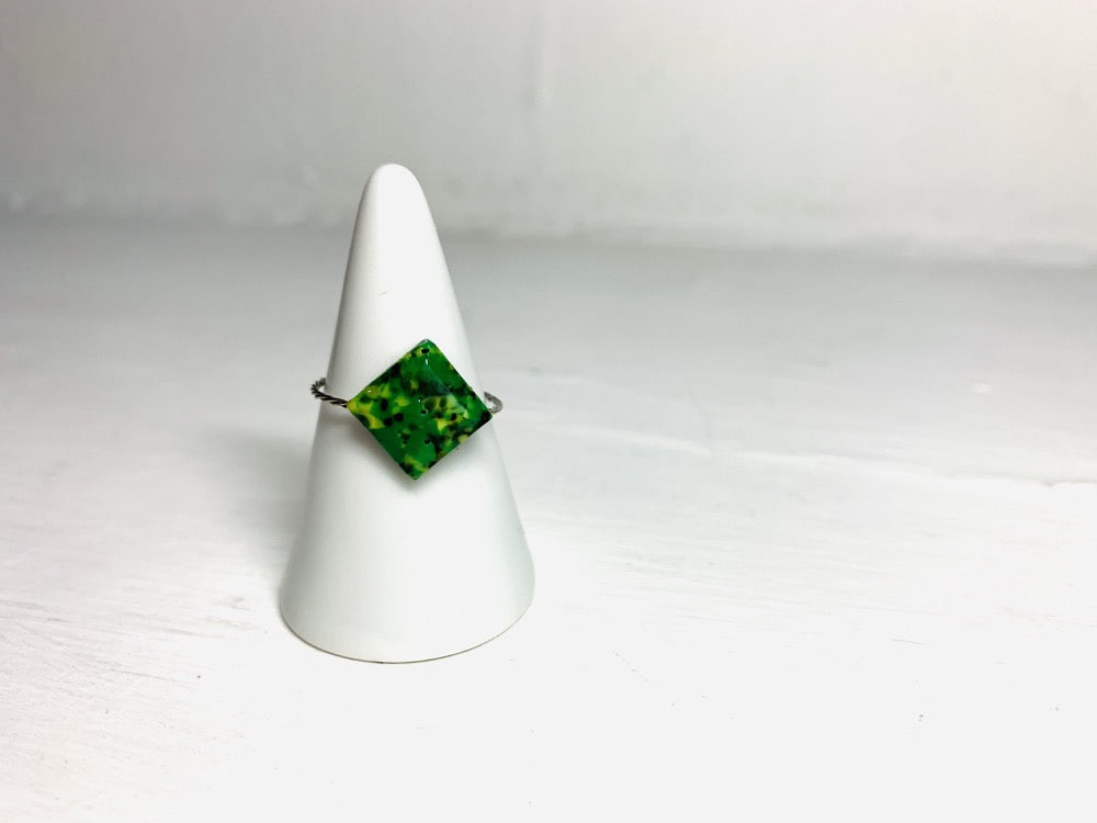 On a white background and slipped on a white cone ring holder is a ring that is cast from recycled 3D prints. The ring is in a faceted square shape with green hues and black colors intermixed to be speckled like granite.