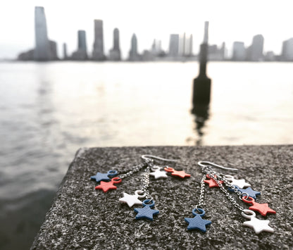 In the background is the Hudson River at dusk, right before  Fourth of July Fireworks. In the foreground, there are two earrings with each with six stars that are red, white, and blue. They are spread out like firework burst. 