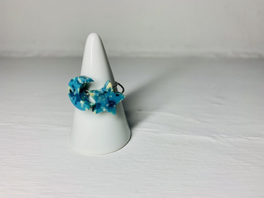 On a white background and slipped on a white cone ring holder are two rings cast from recycled 3D prints. One ring is the shape of a star; the other a present moon. They each have teal, blue, white, and black colors intermixed to be speckled like granite.