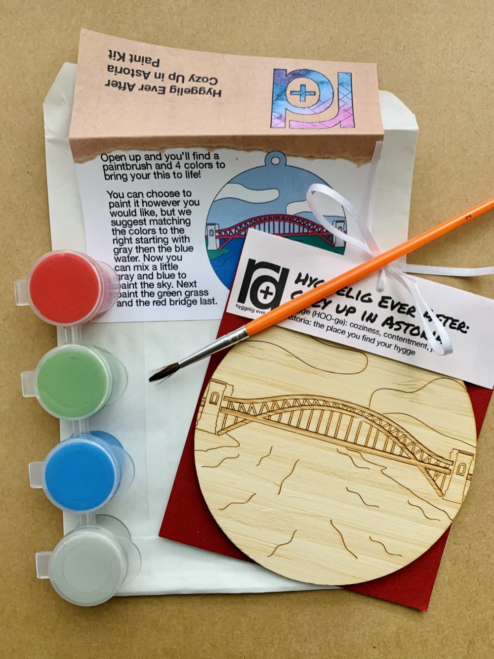 Shown laying on craft paper is the packaging and contents of this DIY Paint Kit. There is a paper envelope with instructions, a paintbrush, a set of 4 paint wells and a wall hanging with a laser cut NYC landmark. 
