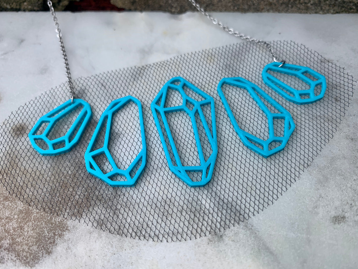 On a white marble background there is a R+D necklace. It is teal eco friendly filament in the shape of five crystals. They don't touch but are embedded with a black tulle fabric to stay together and look like they are floating when being worn. 