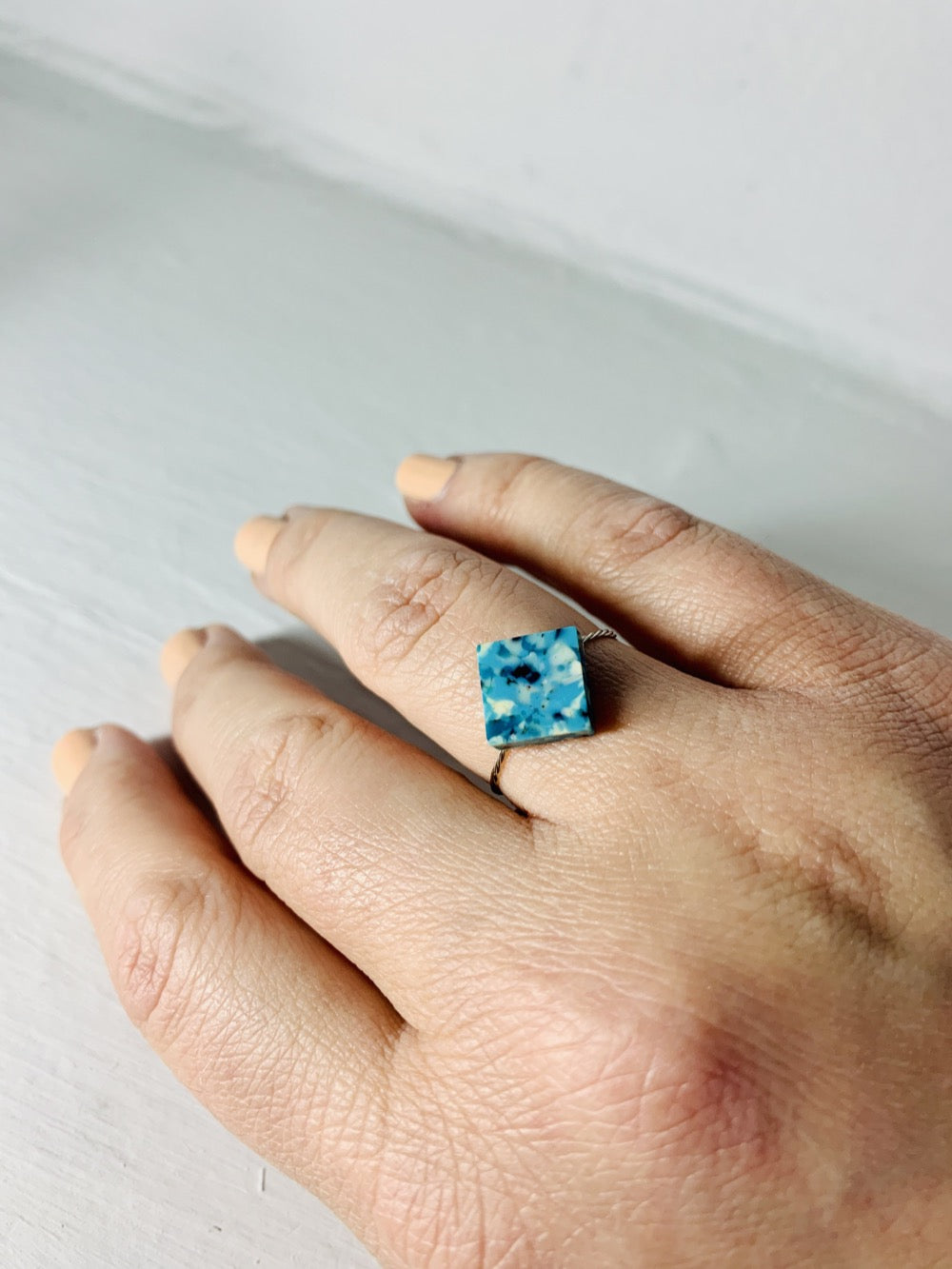 On a white background is someone's hand with a ring on their ring finger. The ring has a thin twisted metal band and a smooth square charm in the middle. The charm is made with recycled 3D prints with teal, blue, white, and black colors intermixed to be speckled like granite.