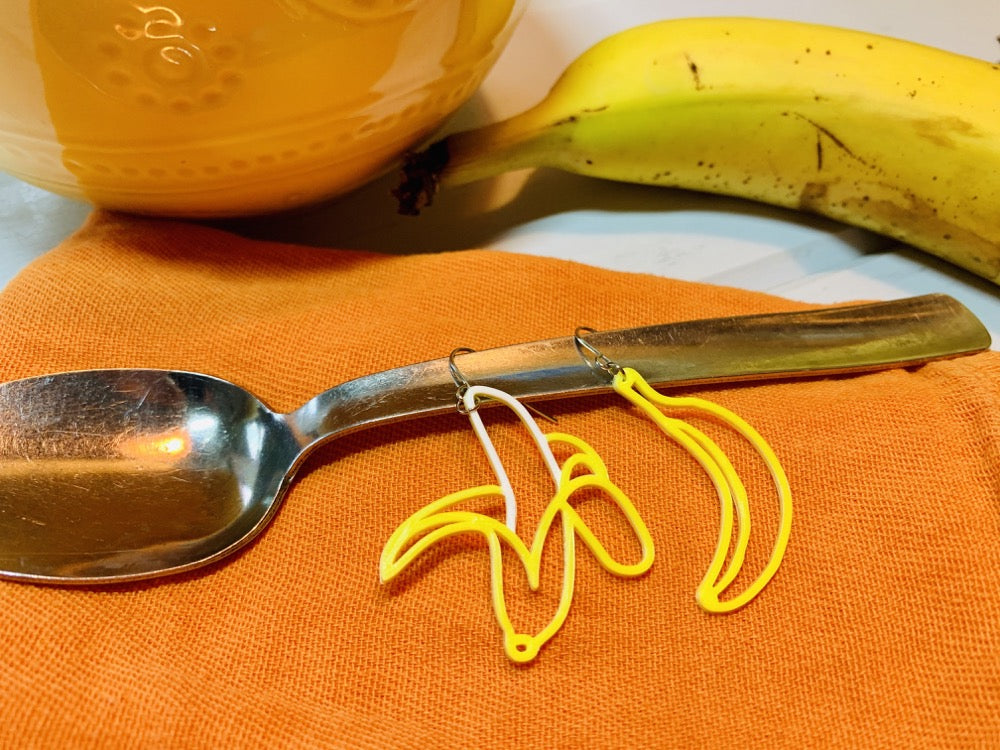 With a banana and a bowl in the background, there is a spoon resting on an orange napkin. on the handle of the spoon are two R+D banana earrings. They are asymmetrical, one is an unpeeled banana and the other is a half peeled banana. They are yellow and white.