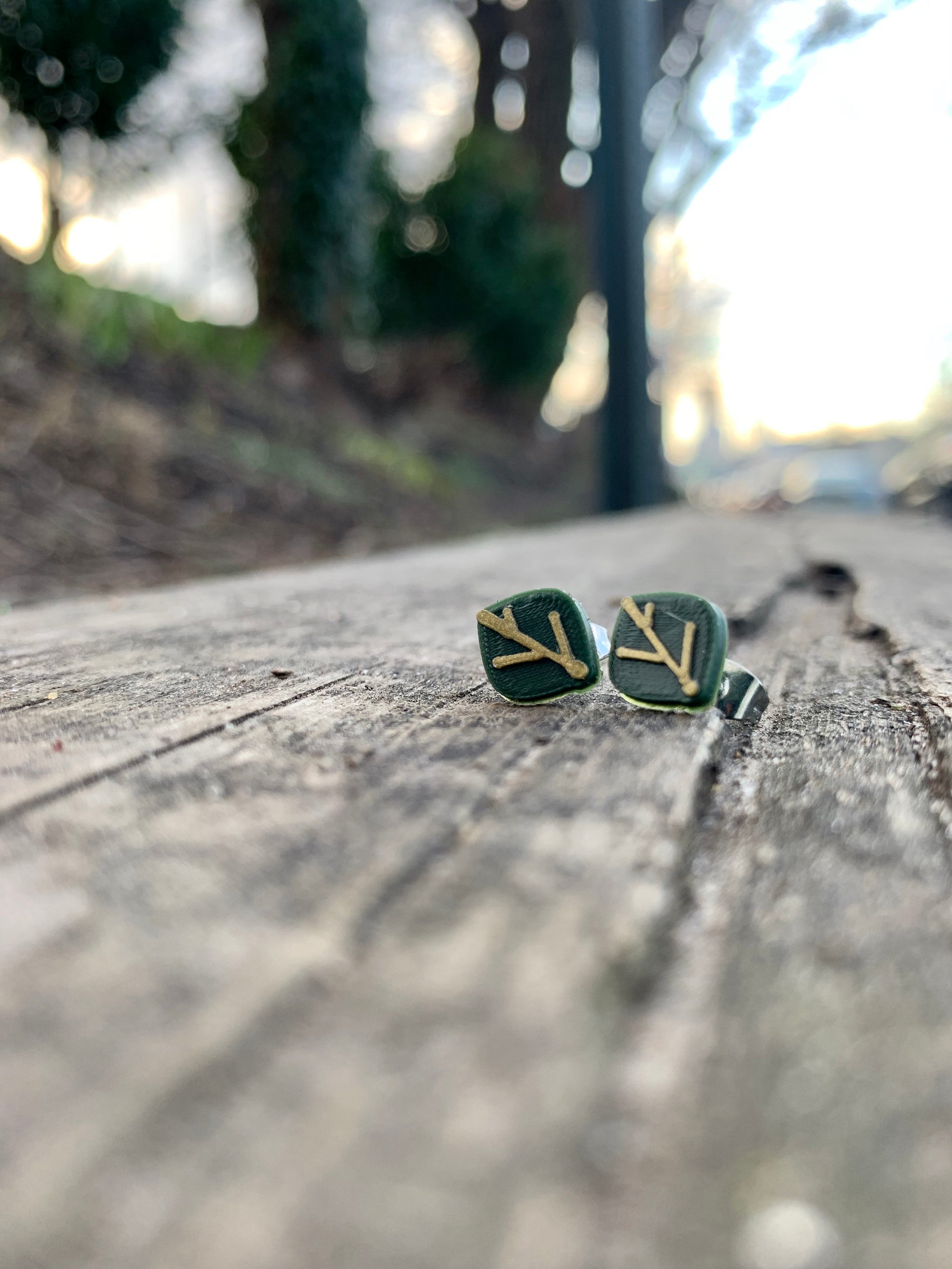 In focus are two R+D earrings shaped like leaves. They are olive green with gold veins. They are resting on weathered wood planks and there is a park blurry in the background.
